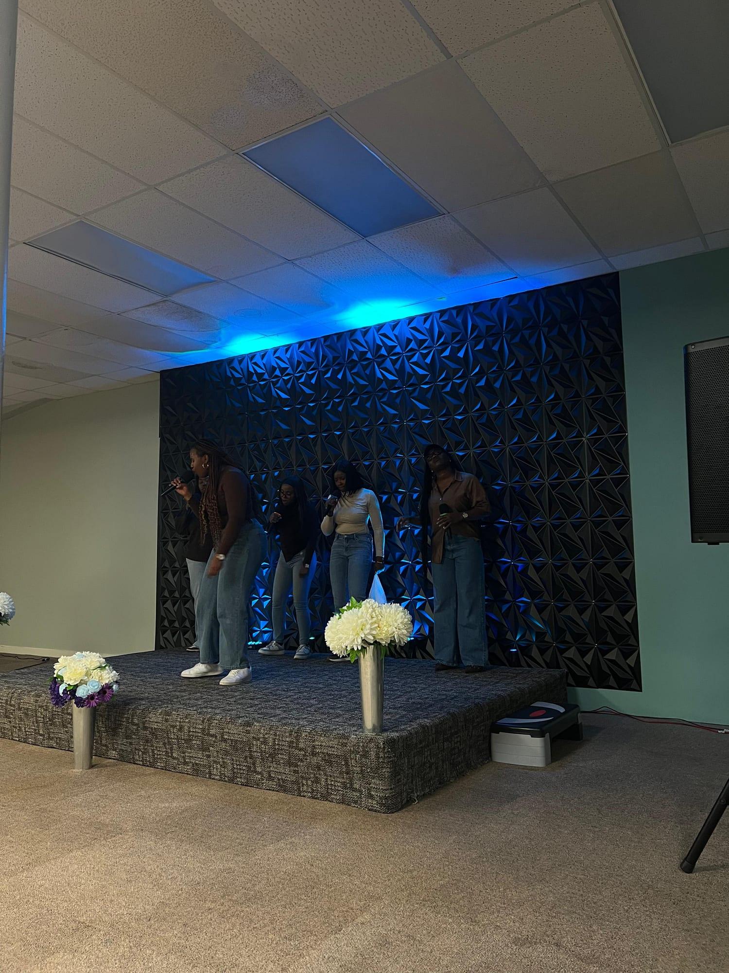 Five singers on the stage in front of a dark patterned wall lit blue, white flowers at the front.