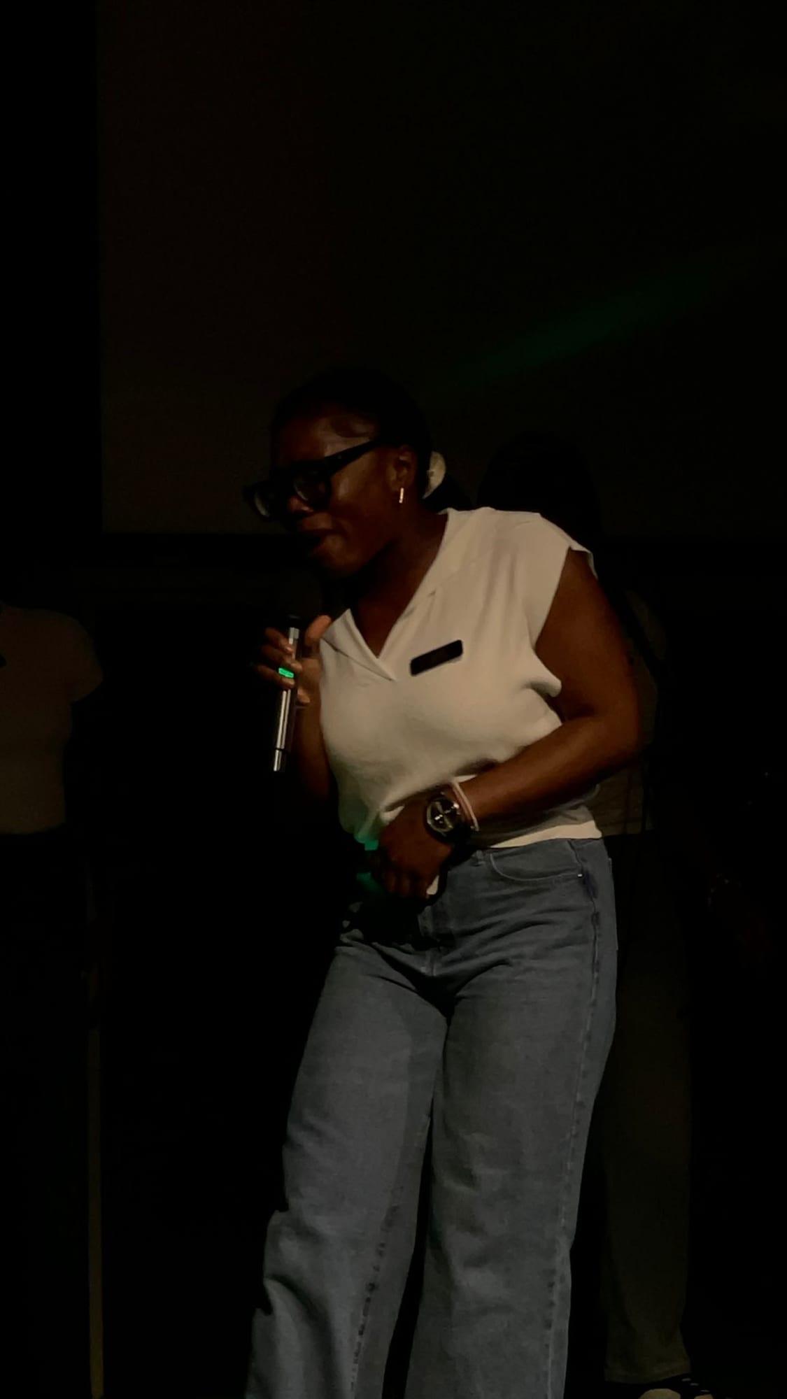 A singer in a white top and glasses leans into the song, microphone in hand, in a dark room.