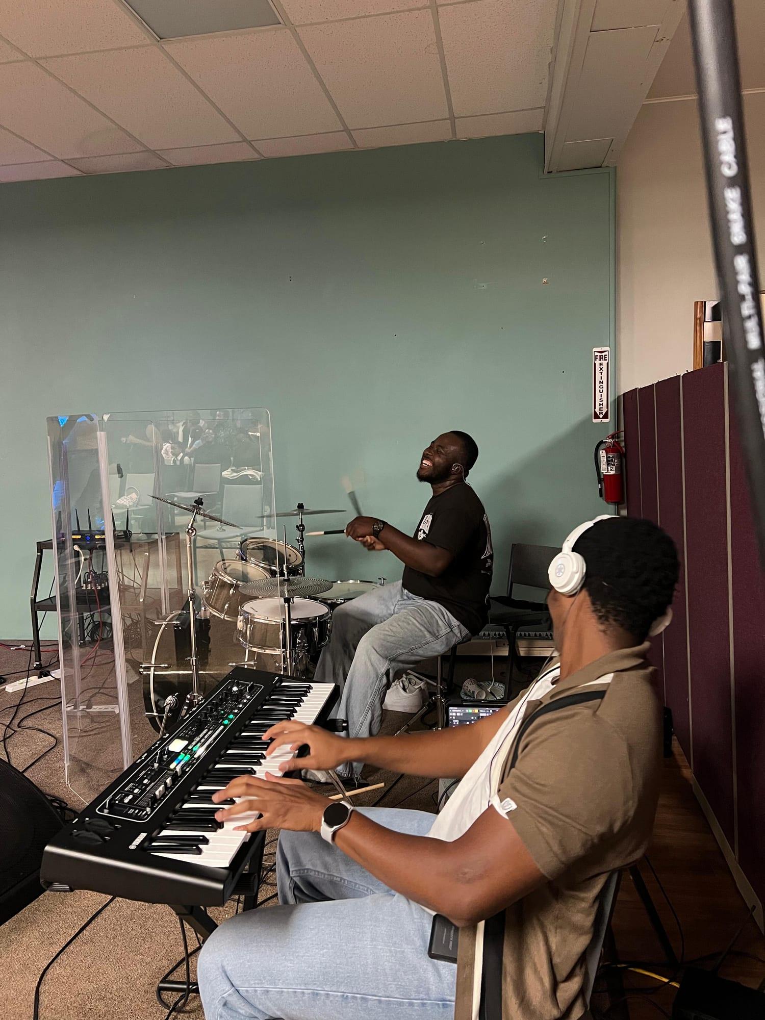 The drummer laughs mid song while the keys player, in white headphones, plays beside him.