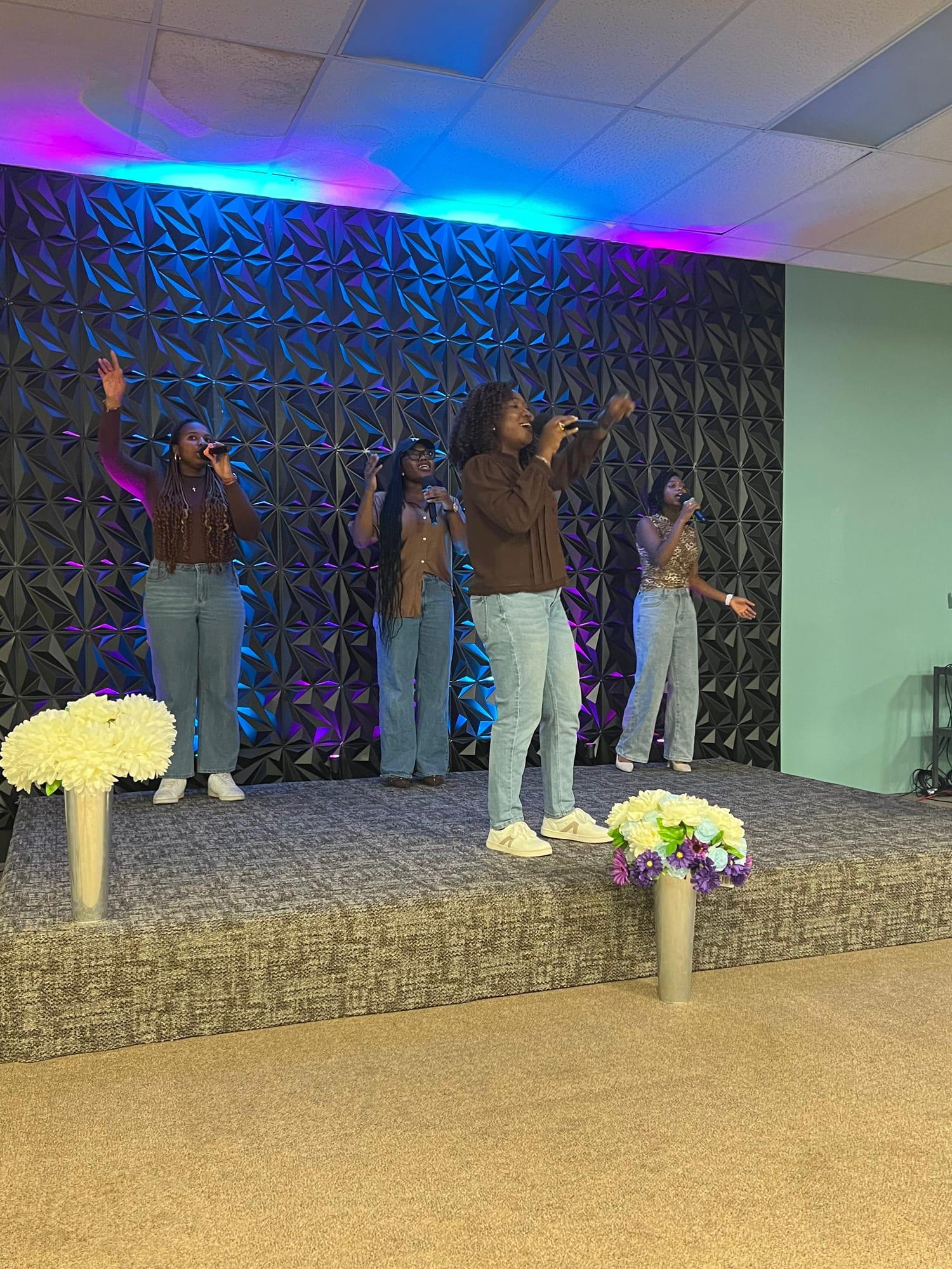 Four singers in brown tops and jeans lead worship on the stage, hands raised, under blue and pink light.