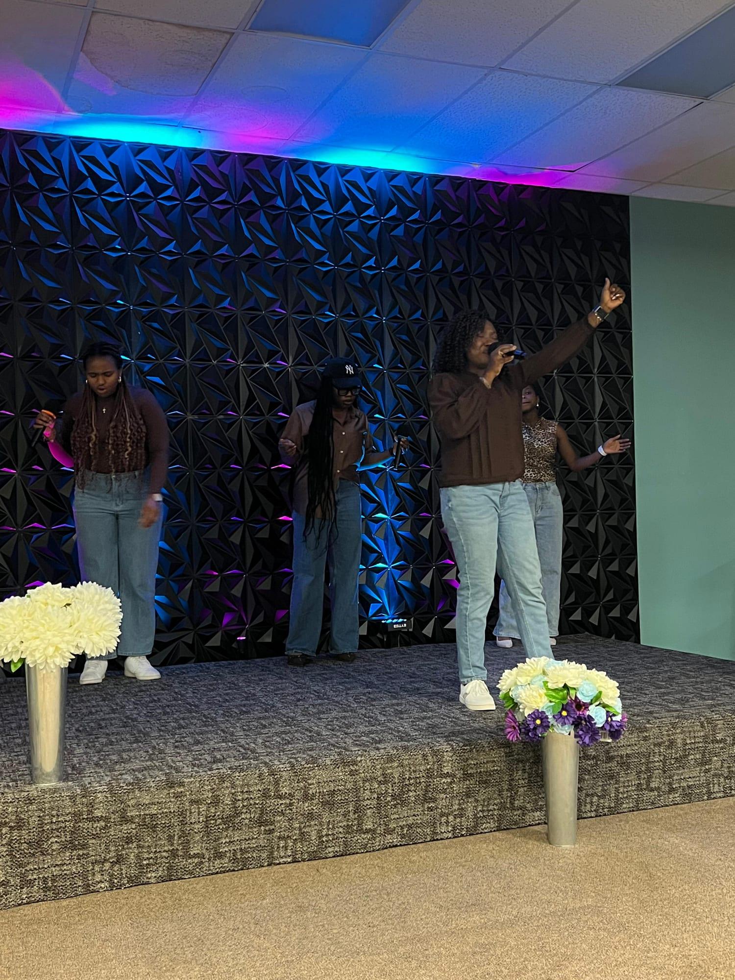 A worship leader lifts one arm high as the team sings behind her on the lit stage.