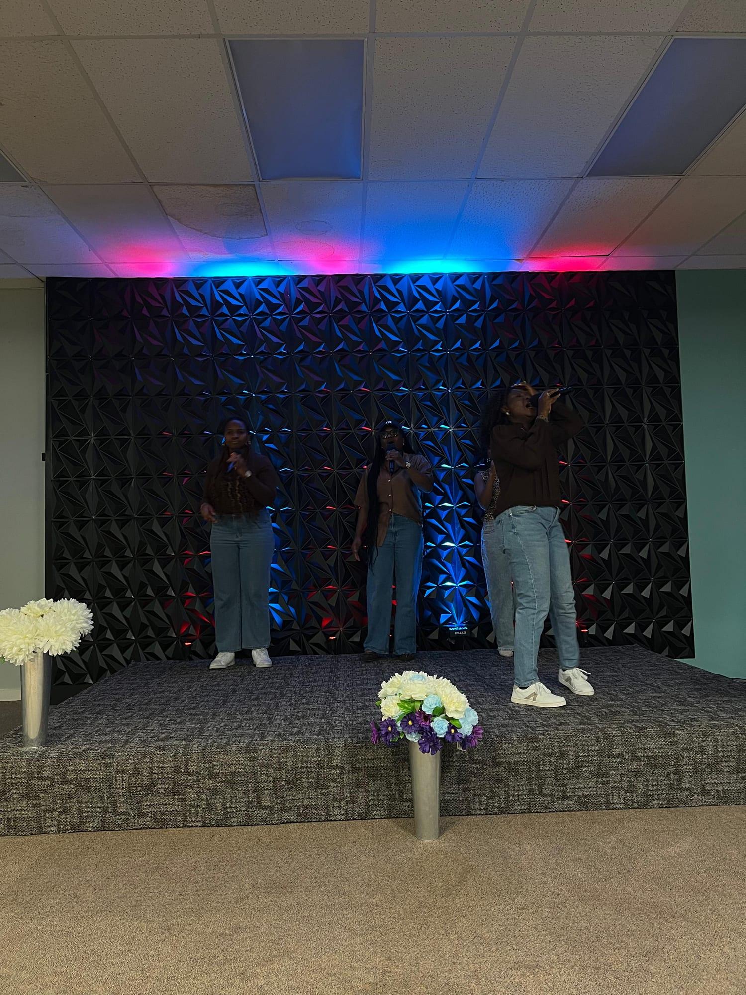 The team sings under red and blue light, the leader holding the microphone close with eyes closed.